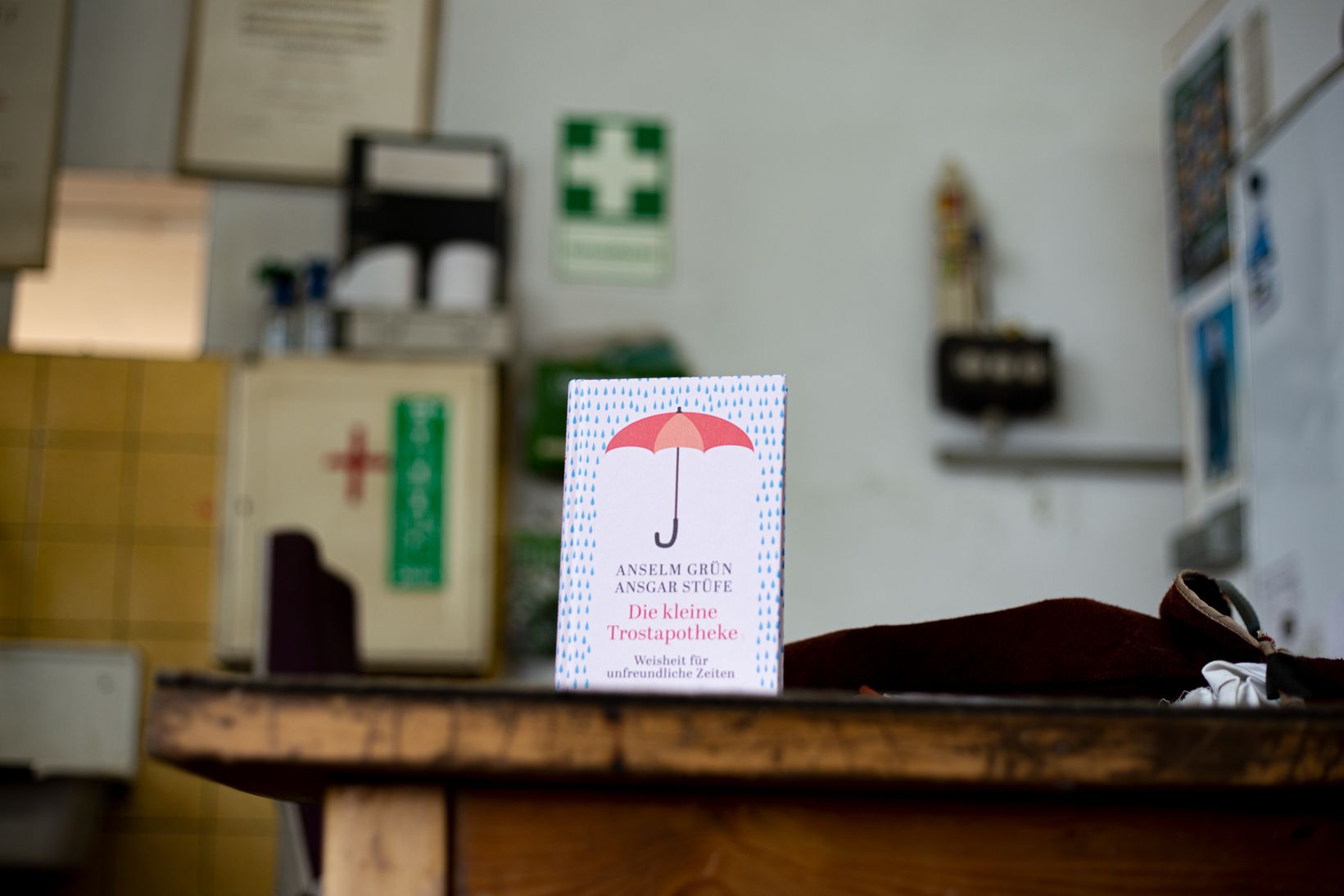 Das Buch "Die kleine Trostapotheke" steht auf einem Schreibtisch, im Hintergrund sieht man einen Erste-Hilfe-Kasten an der Wand hängen
